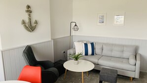 Un salon moderne avec un canapé gris, un fauteuil rouge, une table blanche et un petit pouf gris. Une plante en pot est posée sur la table et une décoration en forme d’ancre orne le mur.