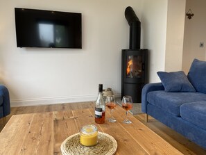 A living room with a flat-screen TV, a wood-burning stove, a blue sofa, and a wooden coffee table with a bottle and glasses.