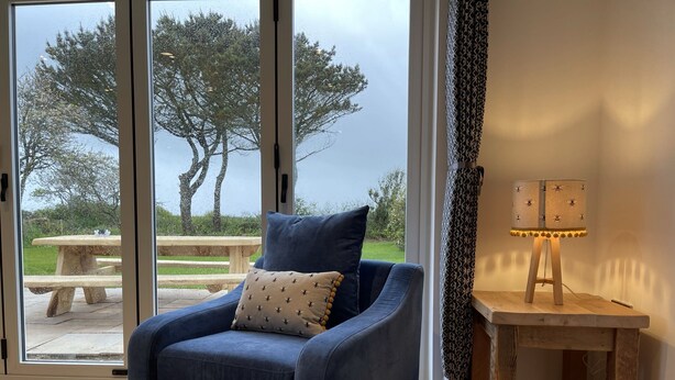 A blue armchair with a patterned pillow, a wooden side table with a lamp, and a view of an outdoor patio with a picnic table and trees. - Holly Cottage @ Arrowan Common Farm (Coverack)