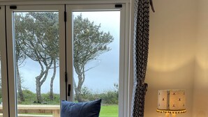 A blue armchair with a patterned pillow, a wooden table with a lamp, and a view of trees through a glass door. - Holly Cottage @ Arrowan Common Farm (Coverack)