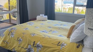 Ein Hotelzimmer mit einem Bett, einem Nachttisch, einer Lampe und Blick auf eine Stadtkulisse durch große Fenster.