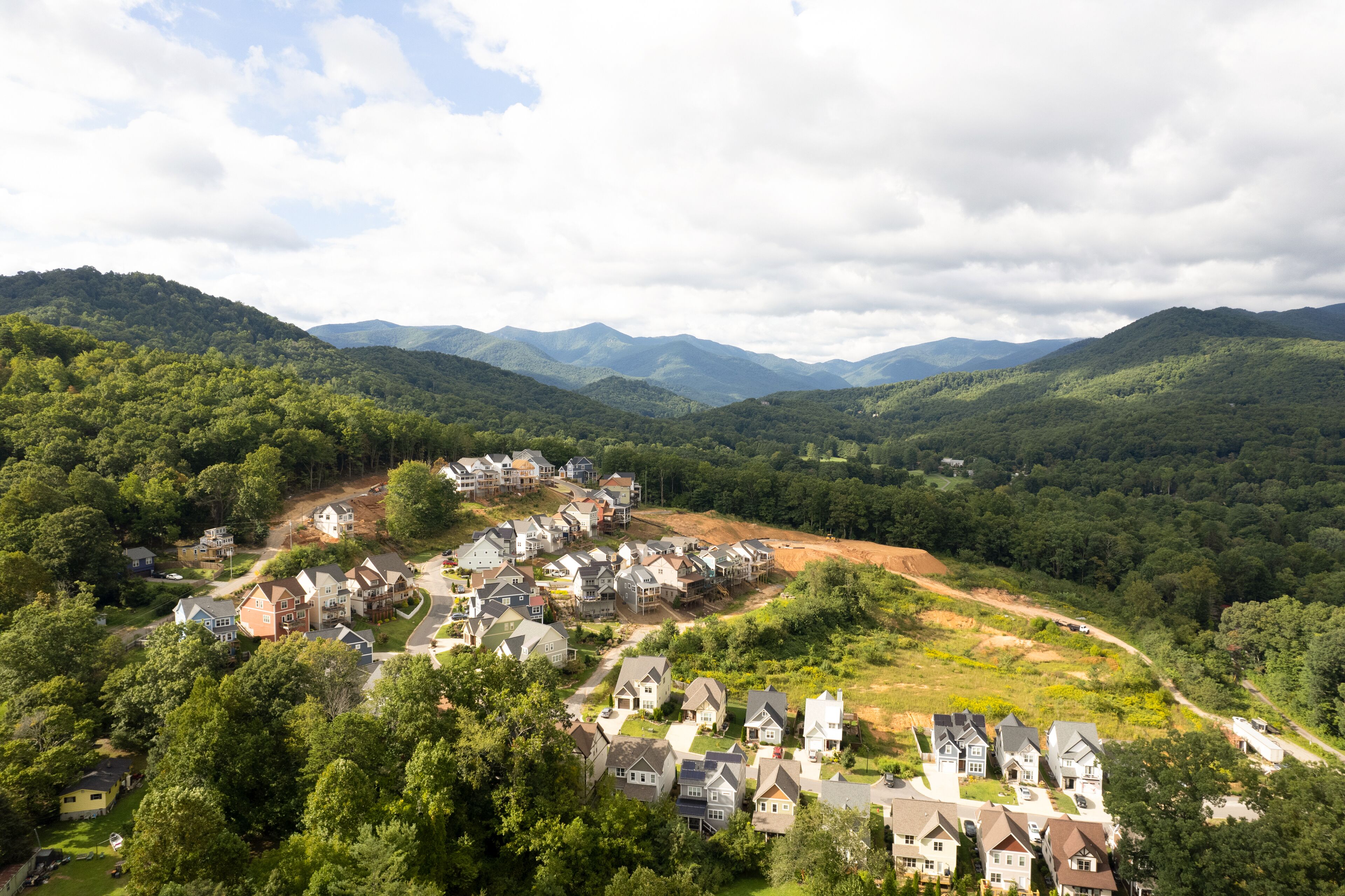 A residential area with houses surrounded by lush greenery and mountains in the background.