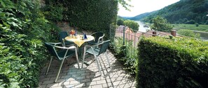 Un patio al aire libre con una mesa y sillas, rodeado de abundante vegetación y vistas a un río y colinas.