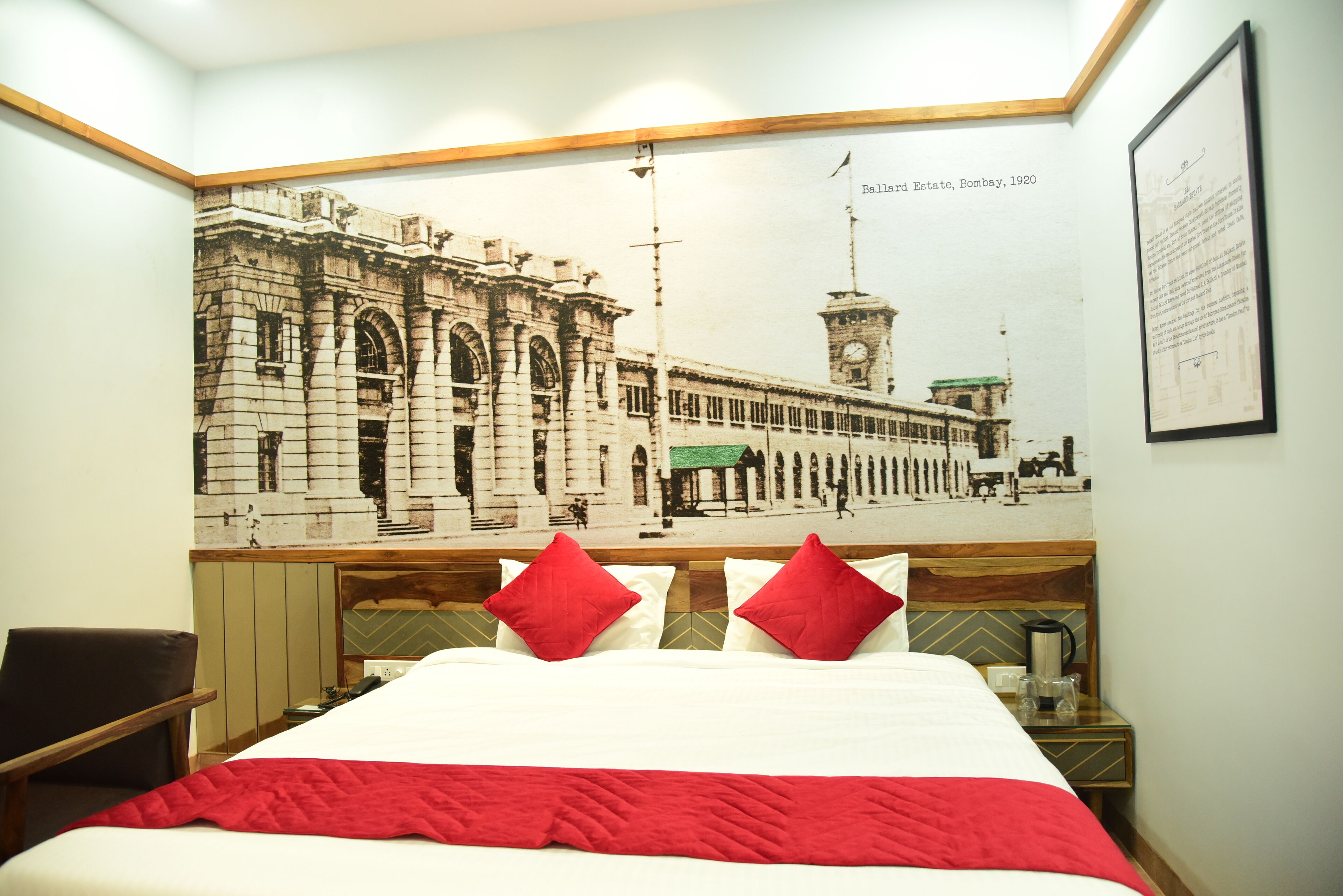 A hotel room with a large bed, two red pillows, a wooden headboard, a nightstand with a coffee pot, and a wall mural of a building.