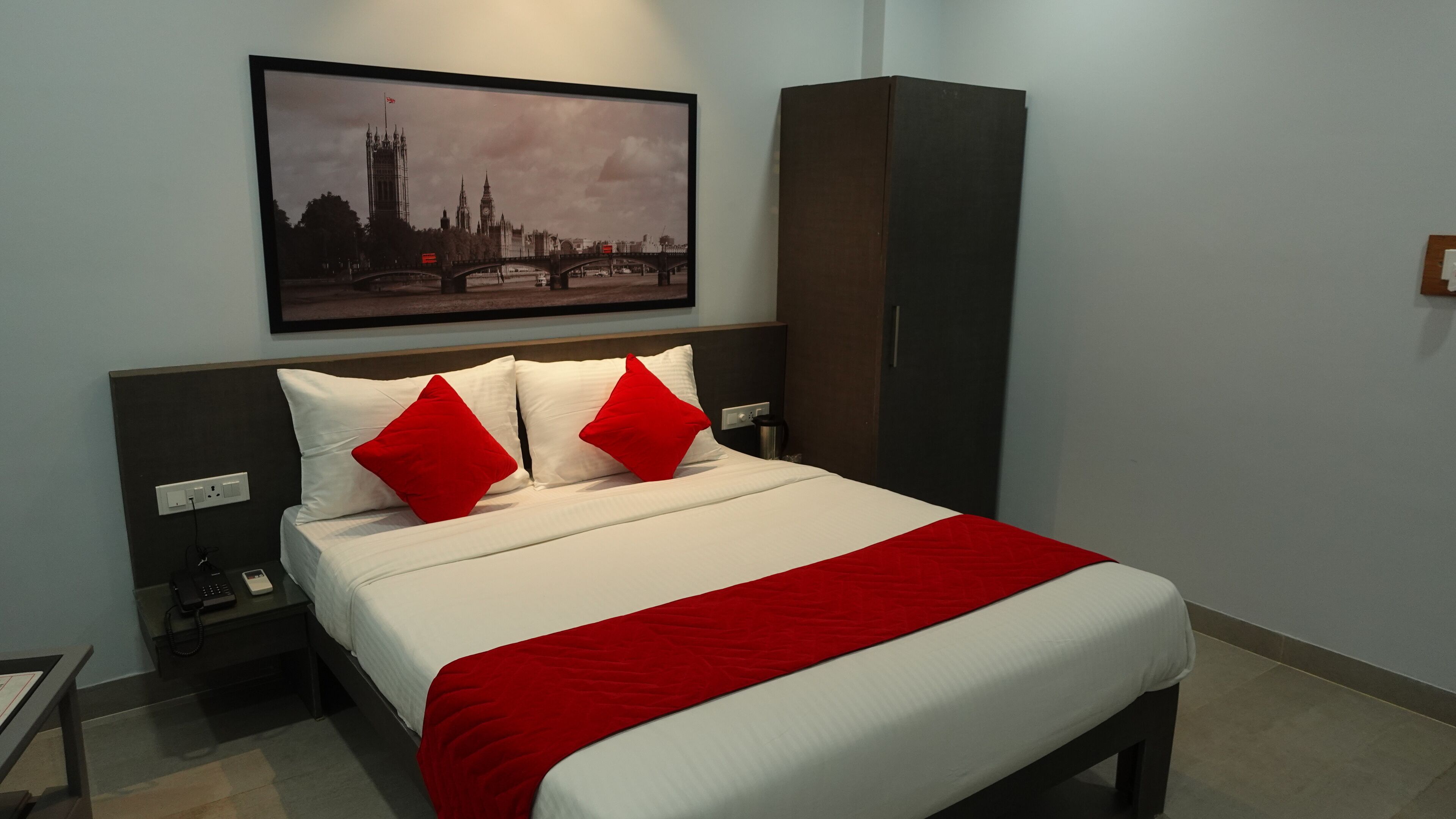 Una camera d'albergo con un letto, un comodino, una testiera e una coperta a due colori, rossa e bianca, e una foto incorniciata di un panorama urbano appesa al muro.