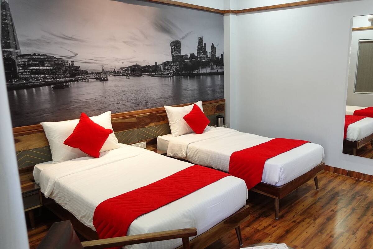 Ein Hotelzimmer mit zwei Betten, jeweils mit weißer Bettwäsche und roten Akzenten, einem großen Spiegel und einem Stadtbild-Wandgemälde.