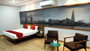 A hotel room with a bed, two chairs, a small table, a nightstand, and a large cityscape painting on the wall. - Annandam Inn-Hotel Anand (Seoni)