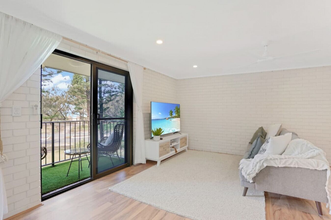 Een moderne woonkamer met een grote schuifdeur naar een balkon, een flatscreen televisie op een witte dressoir, een bank met kussens, en een houten vloer.