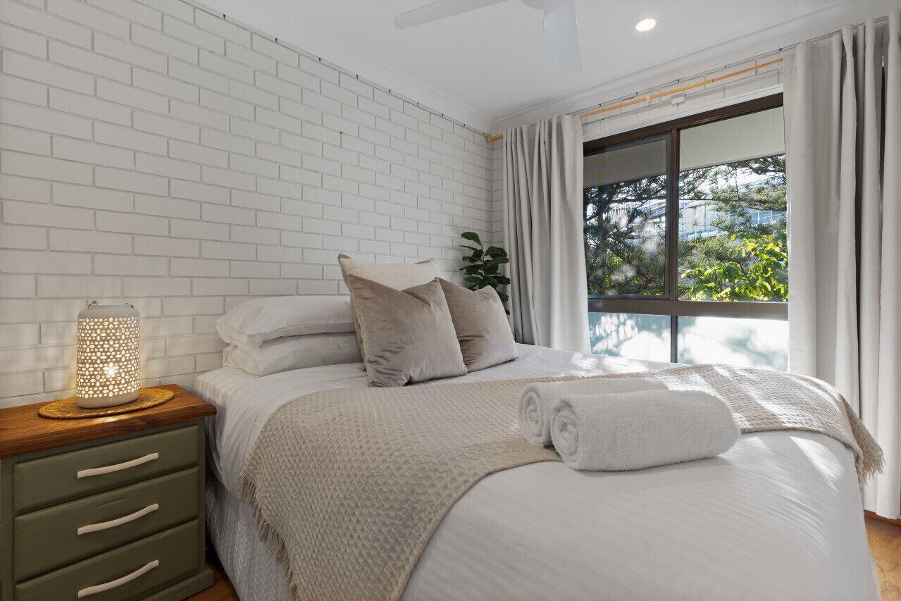 Une chambre moderne avec un mur en briques blanches, une grande fenêtre avec des rideaux, une table de chevet avec une lampe et un lit bien fait, agrémenté d’oreillers et d’une serviette roulée.