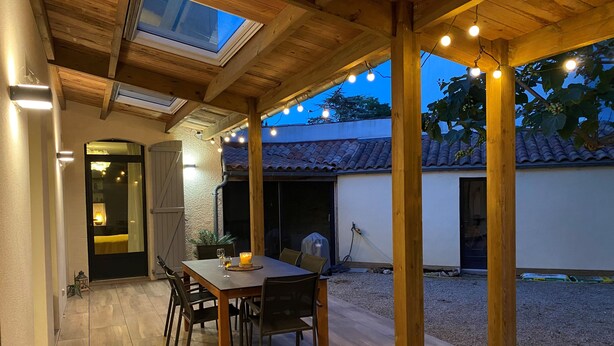 A covered patio with a wooden table and chairs, string lights, and a view of a house with a tiled roof. - Comfortable (122m2) 1.2km from the beach (Jard-sur-Mer)