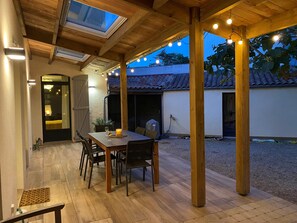 Un porche techado con una mesa y sillas de madera, luces colgantes y vistas a una casa con techo de tejas.