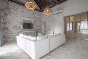 Un salon moderne avec un canapé d’angle blanc, un téléviseur à écran plat fixé à un mur en briques et une grande fenêtre en arc de cercle.