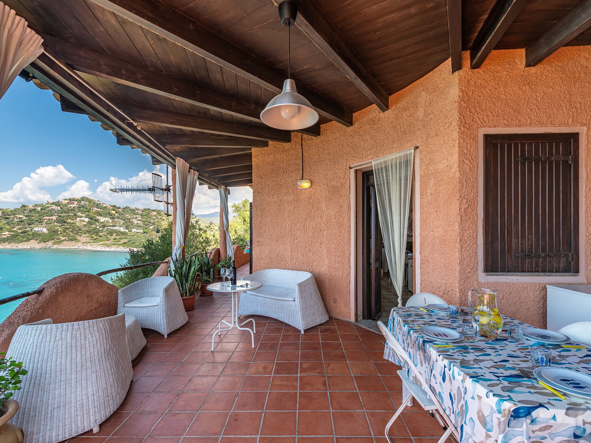 Un patio cubierto con mobiliario de mimbre, una mesa lista para comer y vistas al mar y a las colinas.