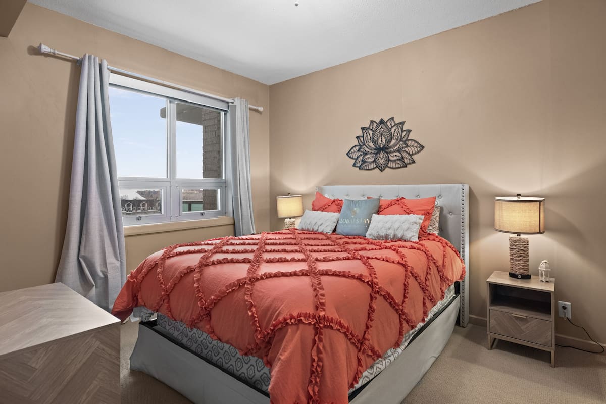 A bedroom with a large bed, a window with curtains, a bedside table with a lamp, and a wall decoration.