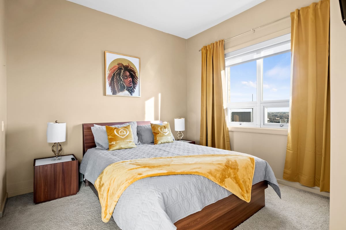 A hotel room with a bed, a nightstand, a window with curtains, and a painting on the wall.