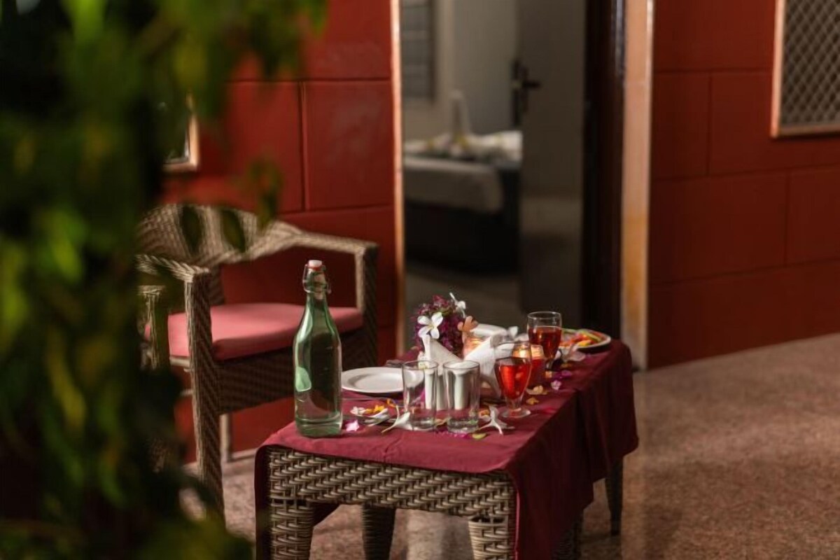 Una mesa de comedor dispuesta con una botella, vasos y una jarrón, con una silla de mimbre al frente.