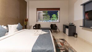 Una habitación de hotel moderna con una cama grande, un escritorio, dos sillas, una pequeña cocina y un ventanal con vista a árboles.
