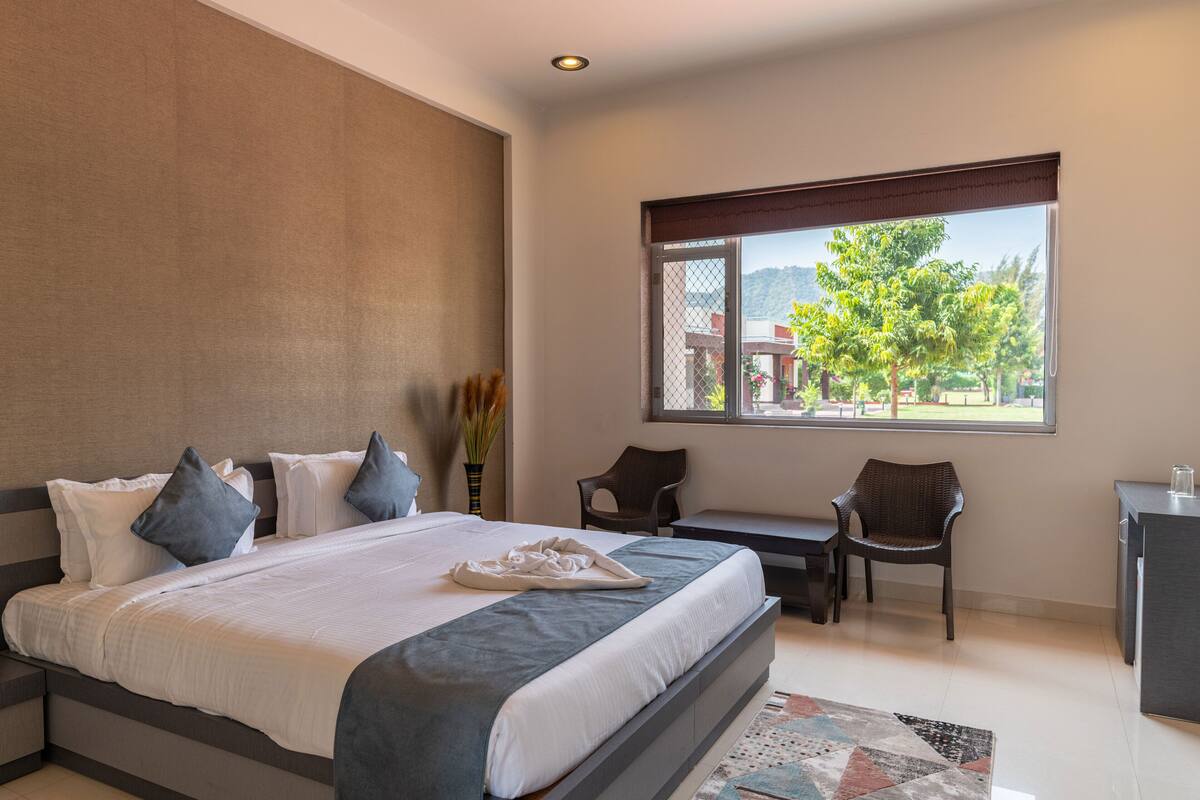 A modern bedroom with a large bed, a desk, and a chair, a window with a view of greenery and buildings, and a small table with a vase of flowers.