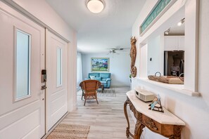 A hallway with a white door, a wall-mounted security device, a ceiling fan, a blue sofa, a wicker chair, a marble console table, and a framed picture on the wall. - Bonita Springs Oasis w/ Wooden Deck & Grill! (Bonita Springs)