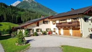 Un bâtiment de style alpin traditionnel, avec une allée en gravier, entouré d’une végétation luxuriante et avec une montagne en toile de fond.