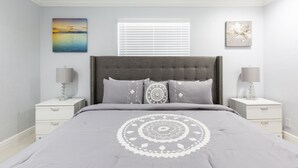 A bedroom with a grey tufted headboard, a grey bedspread with a white circular pattern, two bedside tables with lamps, and a window with blinds.