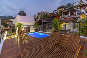 A rooftop pool area with wooden loungers and a small pool, surrounded by plants and overlooking a hillside with buildings.