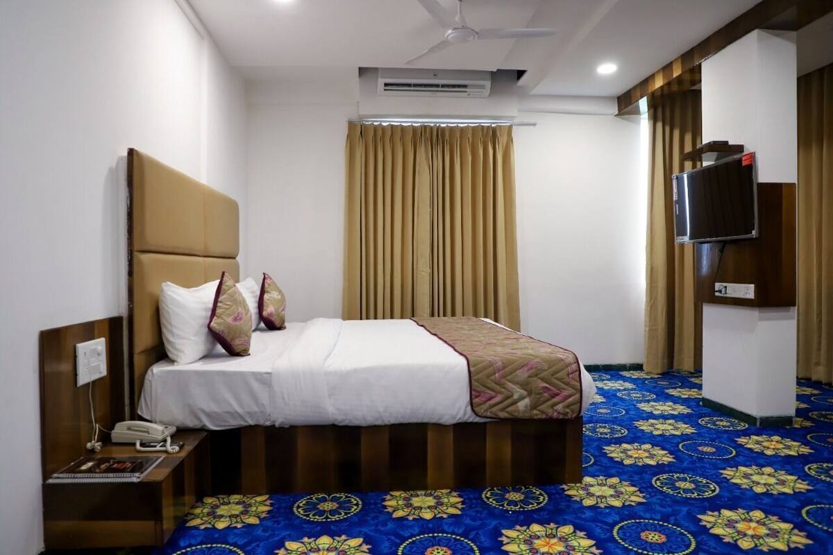 Ein Hotelzimmer mit einem Bett, einem Nachttisch mit Telefon, einem an der Wand befestigten Fernseher und einem gemusterten Teppich.