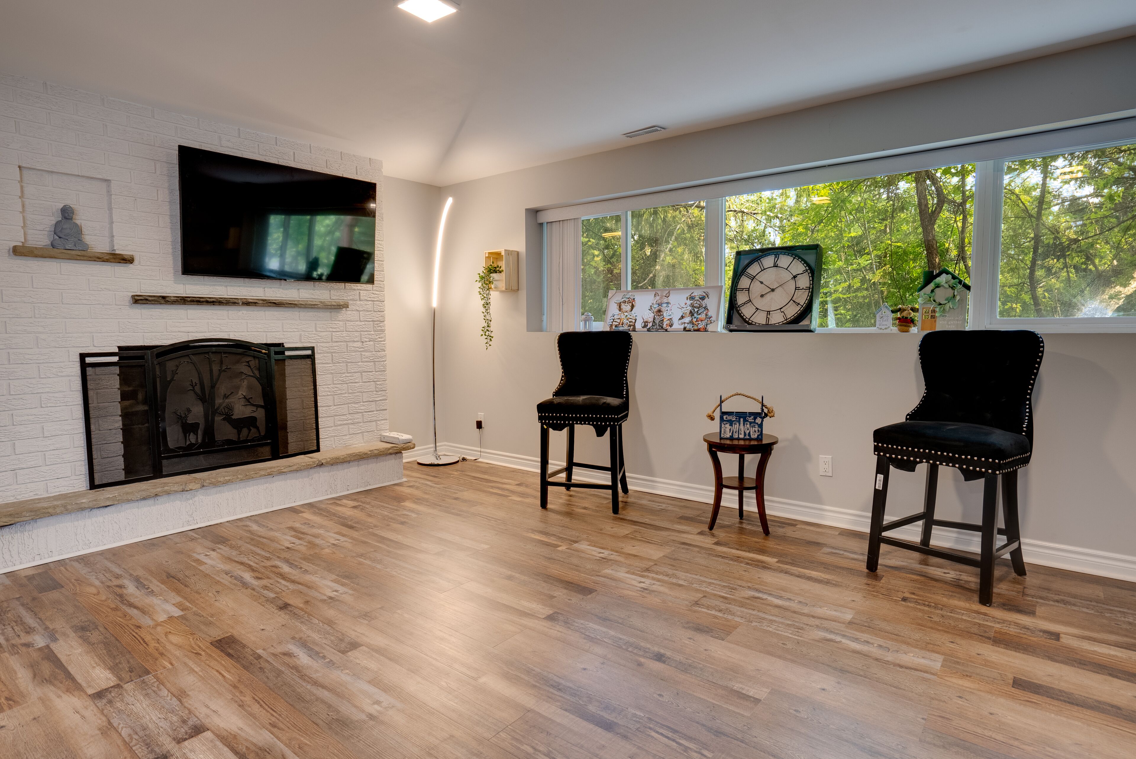 A spacious living room with a fireplace, two black upholstered chairs, a large window with a view of trees, and a flat-screen TV mounted on a white brick wall.