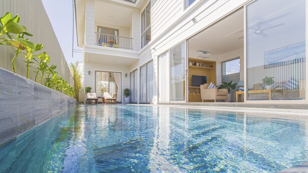 A swimming pool with a clear blue water, surrounded by a white building with balconies and sliding glass doors. - The Manor Hoi An Villa (Da Nang)