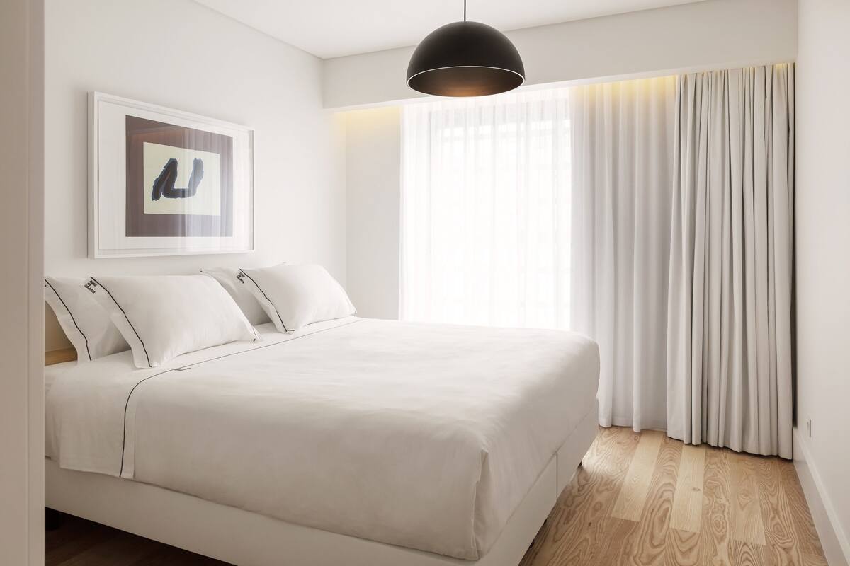 A modern bedroom with a large bed, a pendant light, a framed artwork, and sheer curtains.