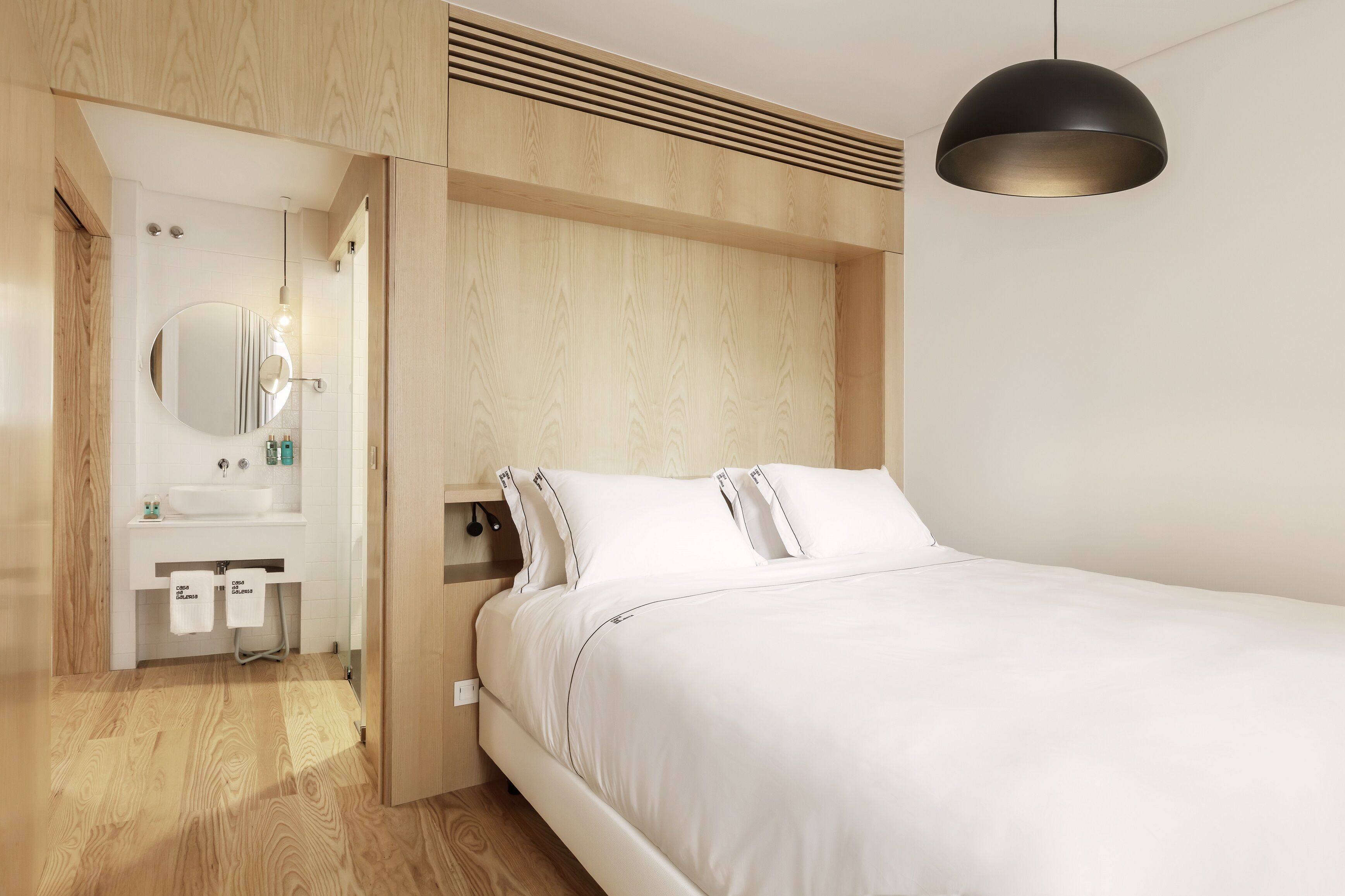 A modern bedroom with a white bed, a round mirror, and a white sink in the adjacent bathroom.