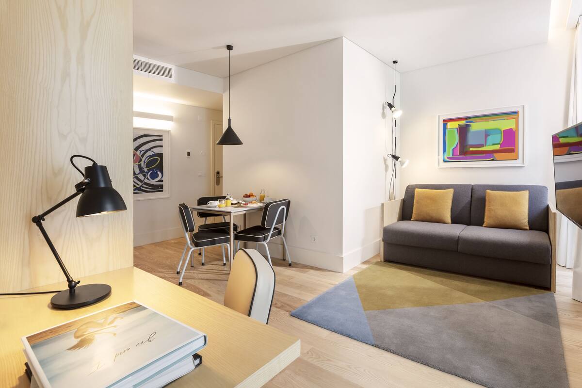 A modern living room with a sofa, a dining table with chairs, a desk with a lamp, and a painting on the wall.