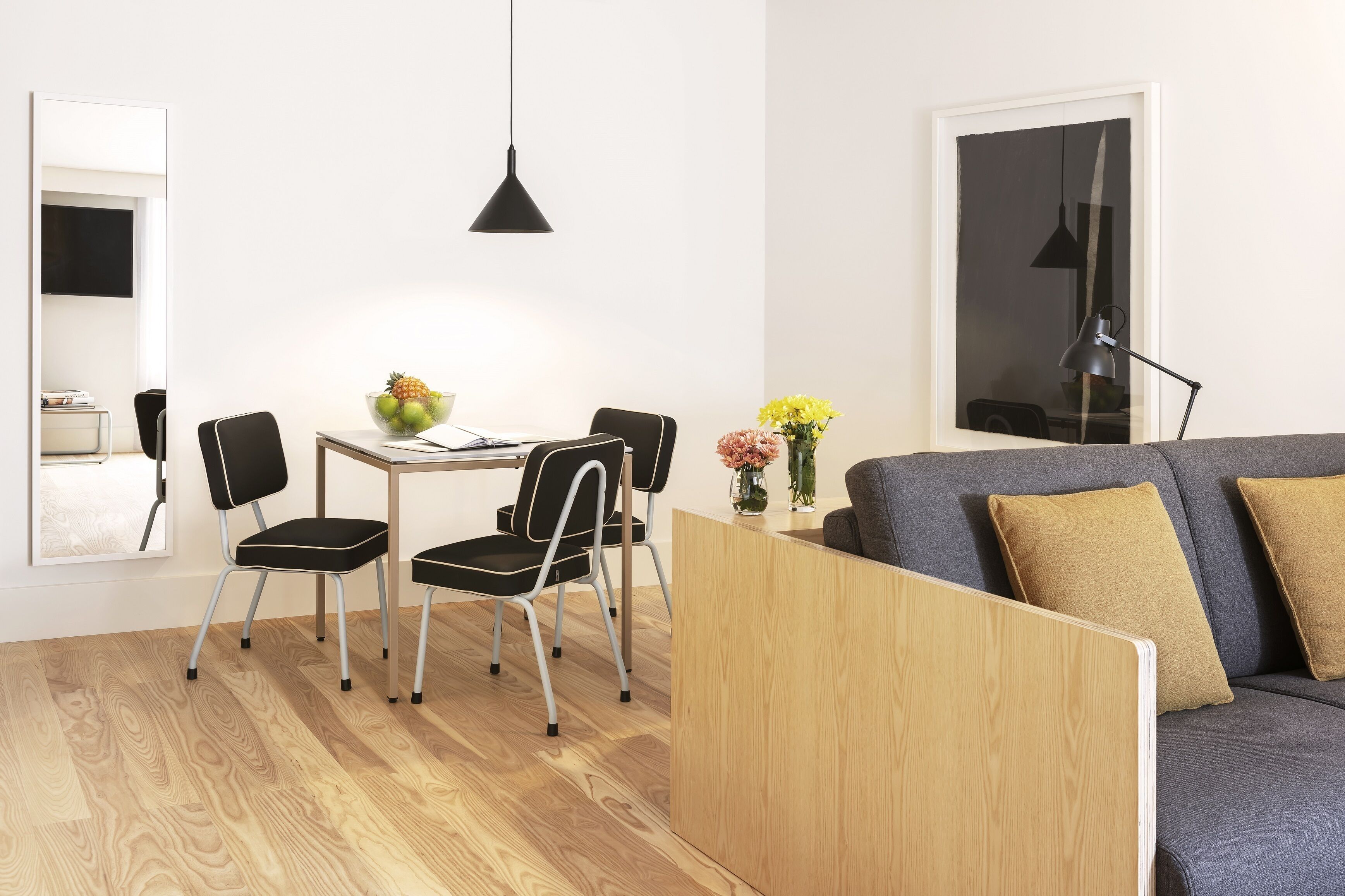 A modern living room with a wooden floor, a gray sofa with two cushions, a dining table with chairs, a black pendant light, and a vase with flowers.
