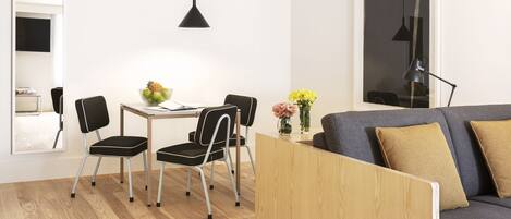 A modern living room with a wooden floor, a gray sofa with two cushions, a dining table with chairs, a black pendant light, and a vase with flowers.
