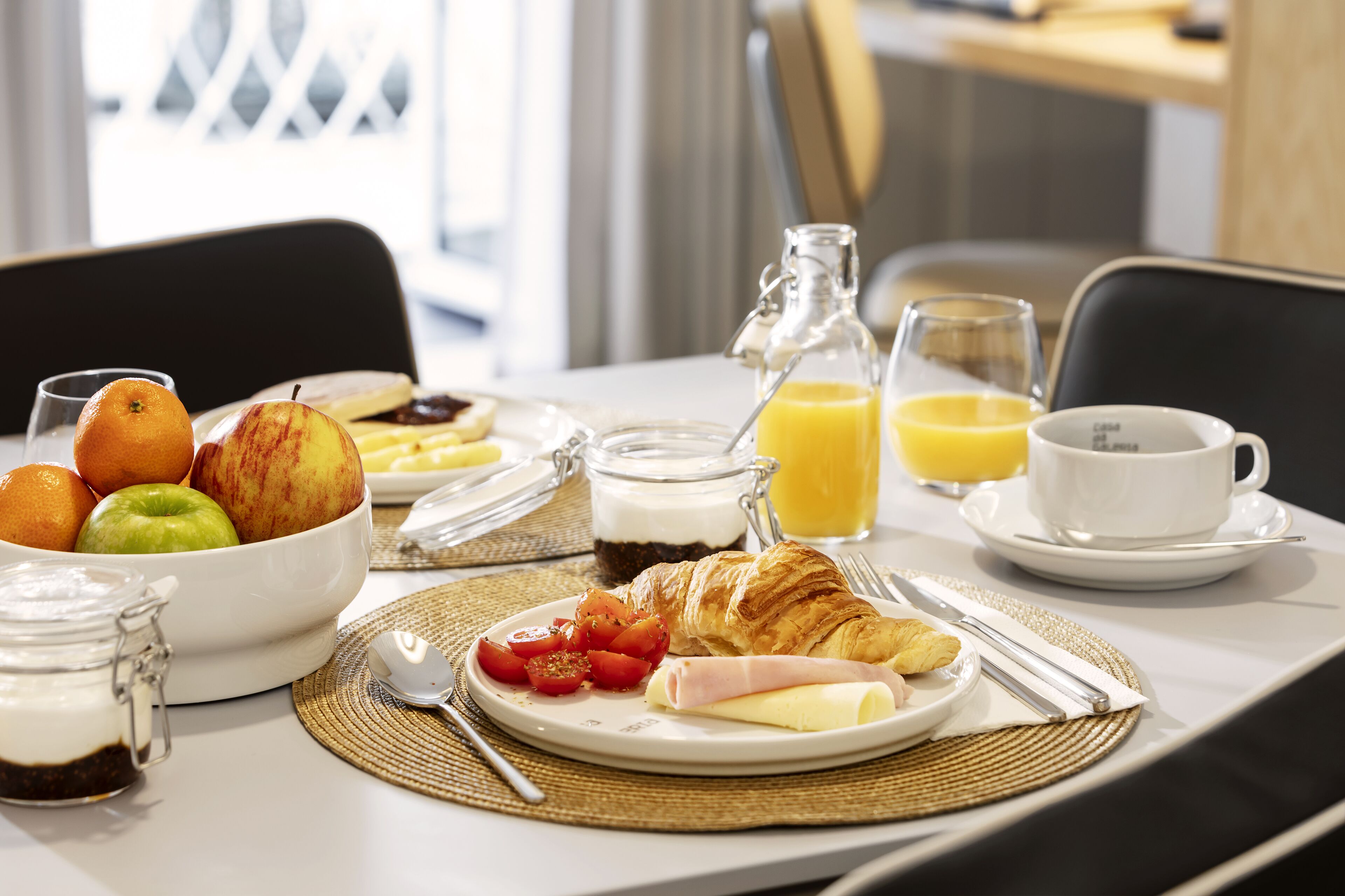 A breakfast table with croissants, tomatoes, cheese, ham, and a bowl of fruit.