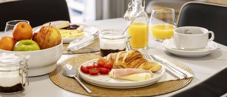 A breakfast table with croissants, tomatoes, cheese, ham, and a bowl of fruit.