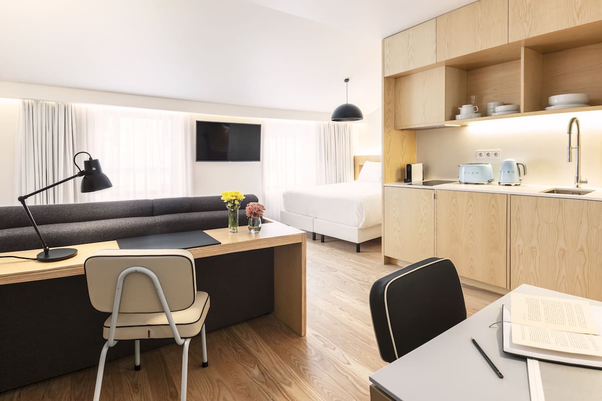 A modern hotel room with a sofa, desk, chair, and a small kitchenette.