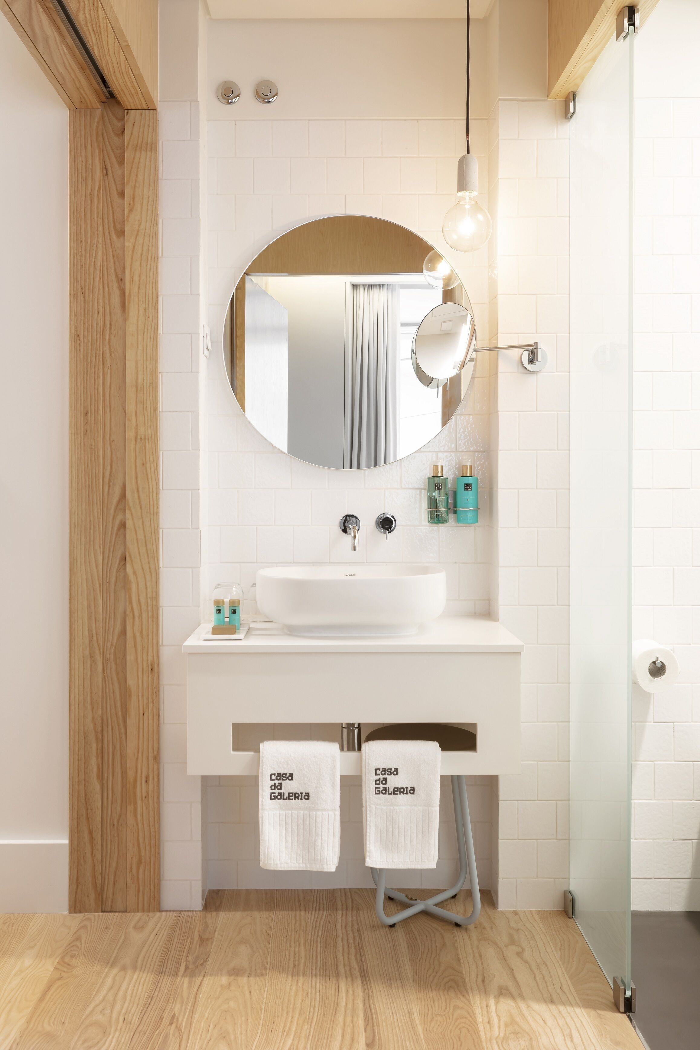 A modern bathroom with a wooden floor, white tiles, a round mirror, a white sink, and two towels hanging on a rack.
