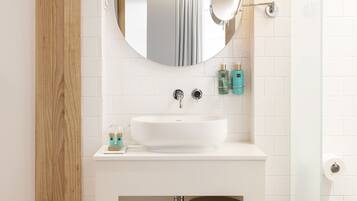 A modern bathroom with a wooden floor, white tiles, a round mirror, a white sink, and two towels hanging on a rack.