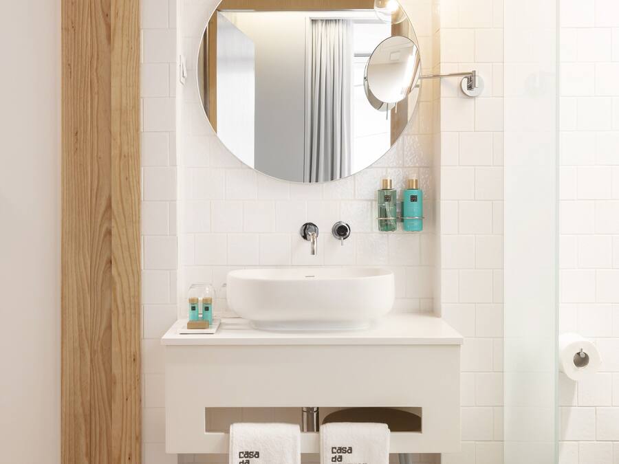 A modern bathroom with a wooden floor, white tiles, a round mirror, a white sink, and two towels hanging on a rack.