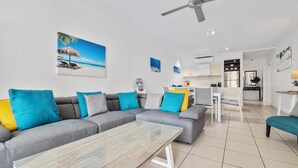 A modern living room with a grey sectional sofa, a glass coffee table, and a ceiling fan. There is a kitchen area in the background with white cabinets and a dining table.