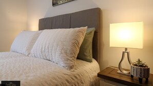 Ein ordentlich gemachtes Bett mit grauem Kopfteil, einer Nachttischlampe und einem kleinen Topfpflanz auf dem Nachttisch.