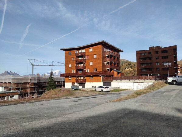 Un immeuble à plusieurs étages doté de balcons, d’un espace de stationnement et ayant une chaîne de montagnes en arrière-plan.
