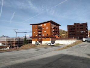 A multi-story building with balconies, a parking area, and a mountainous backdrop.