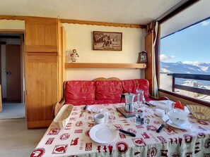 Una habitación acogedora con una mesa de comedor para dos, un aparador de madera, un sofá rojo y un ventanal con vista a la montaña.