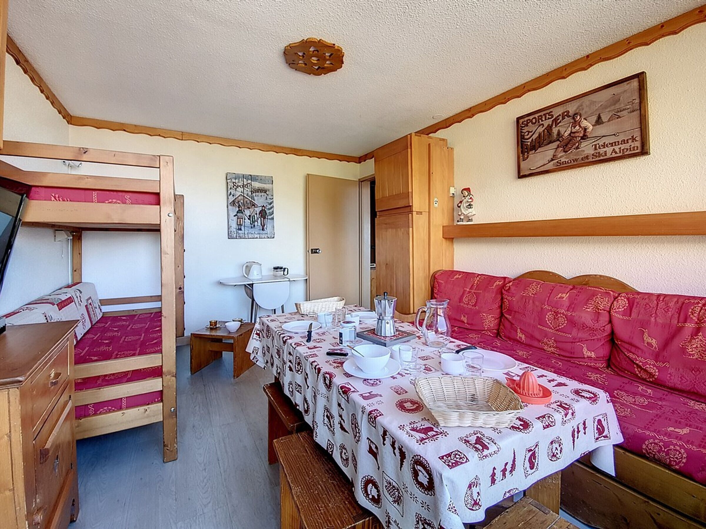 Une pièce chaleureuse avec des lits superposés, une table à manger dressée avec des assiettes, des tasses et un pichet, et un canapé rouge.