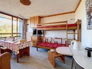 Um quarto aconchegante com uma cama beliche, uma pequena escrivaninha com um computador, uma mesa com toalha e uma janela com vista para árvores.