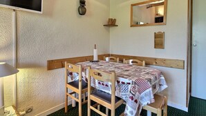 Ein Essbereich mit einem Tisch für vier Personen, Holzsesseln, einem an der Wand montierten Fernseher, einer Deckenleuchte und einem Spiegel über dem Tisch.