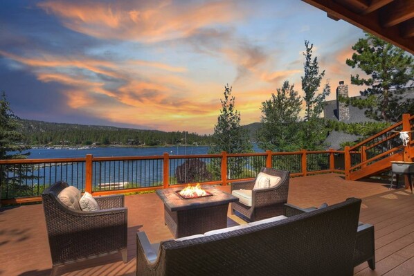 A deck with wicker furniture and a fire pit, overlooking a lake and forested hills.