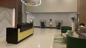 Un espace d’accueil moderne avec un comptoir en marbre, des fauteuils à revêtement vert, une petite table agrémentée d’une plante et un luminaire circulaire original.